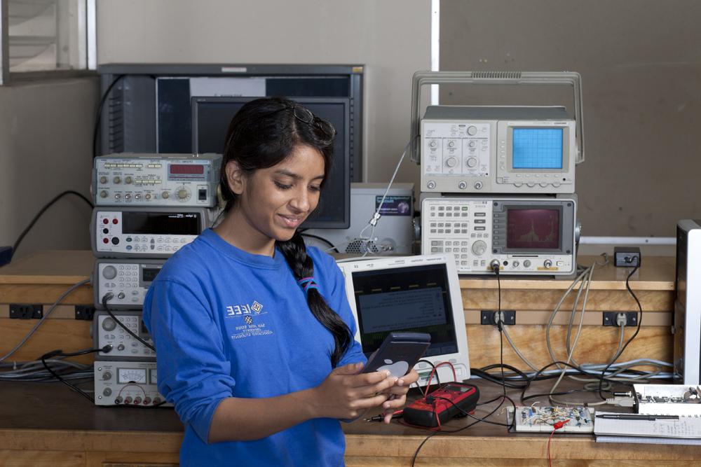 一个穿蓝色衬衫的学生在实验室里，她身后有许多电子设备.
